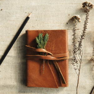 a leather bound journal sits on a beige fabric background with two dried flowers and a pen ready to write.