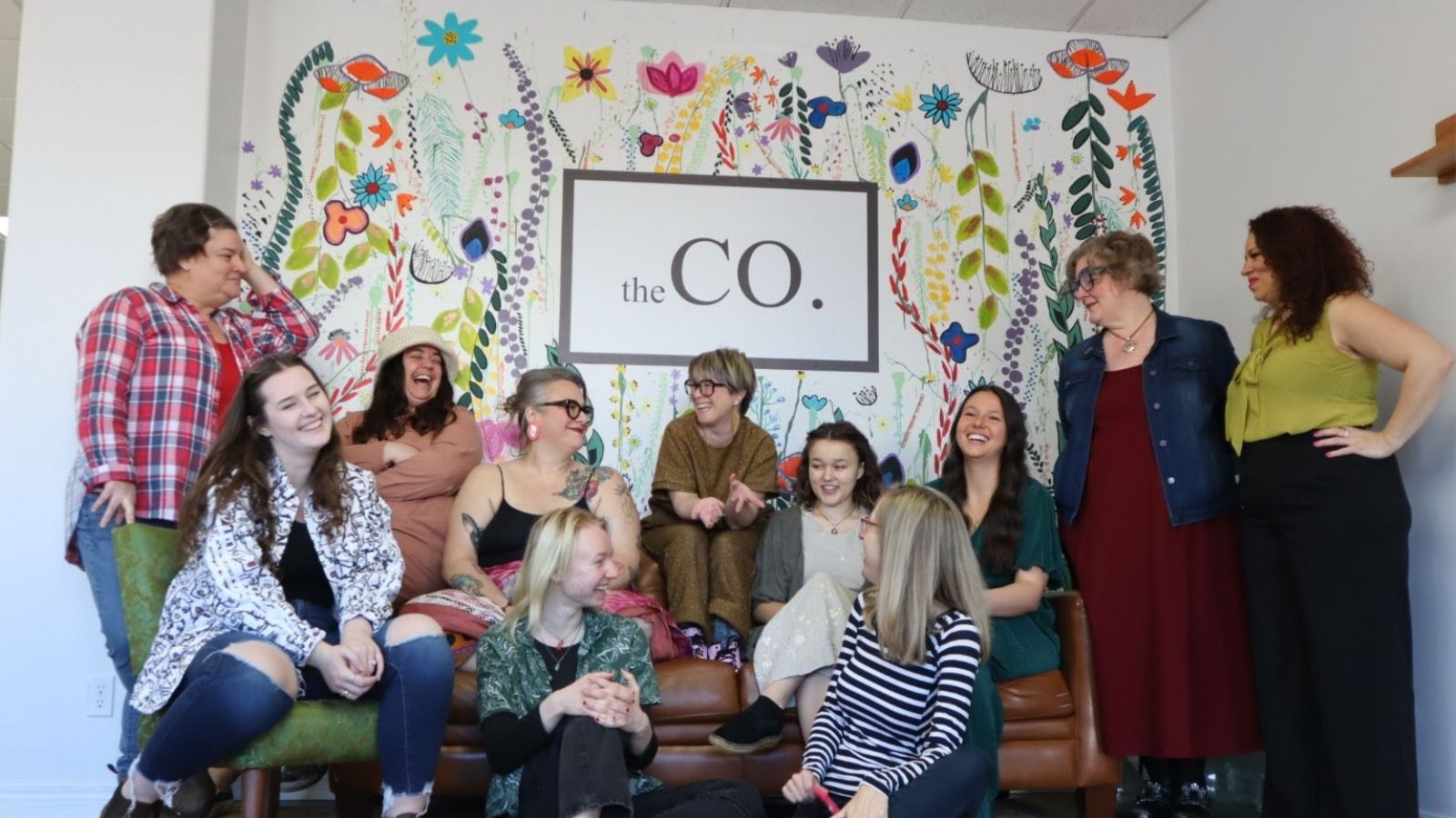 Women stand in front of a wall painted with colourful flowers and the words "the CO."