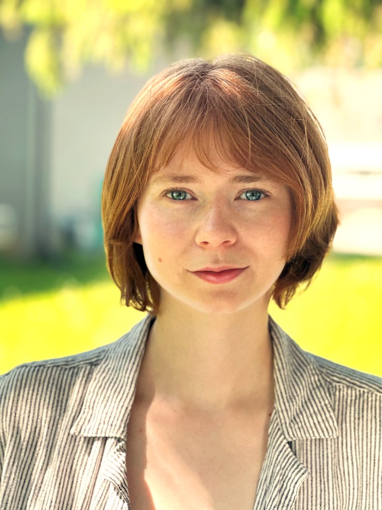 A photo of illustrator Brooklin Stormie, with chin-length strawberry blond hair, against a green grassy background.